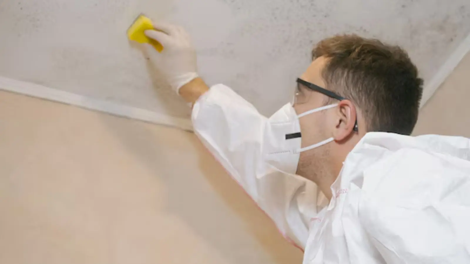 Technician in a mask and protective suit wiping a mold-stained wall